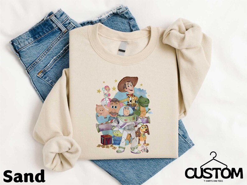 A beige t-shirt with a cartoon character design, including a cowboy hat, a dinosaur, and other characters, is displayed on a white background.