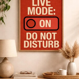 Può includere: Una stampa rossa incorniciata con le parole "LIVE MODE: ON DO NOT DISTURB" in bianco. La stampa è esposta su una mensola di legno con una lampada, libri, occhiali e un vassoio decorativo con una tazza e un vaso.