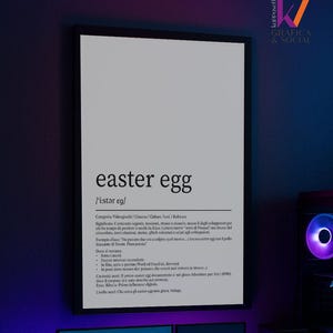 Può includere: Un poster incorniciato con la parola "easter egg" in grassetto. Sotto, una definizione ed esempi in italiano. Il poster è esposto sopra una scrivania con due monitor per computer, altoparlanti e una tastiera.