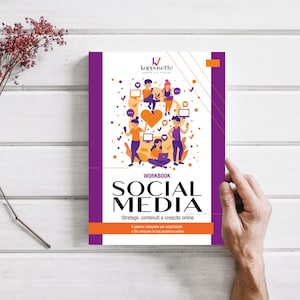 Peut inclure: Un cahier intitulé "Social Media" avec une couverture violette et orange. La couverture présente une illustration de personnes interagissant avec des icônes de médias sociaux. Le livre est tenu par une main sur une surface en bois blanc.