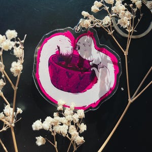 May include: A clear acrylic keychain featuring a white cat drinking from a magenta bowl. The cat has a long tail and is set against a white background with a magenta outline. A silver keyring is attached.