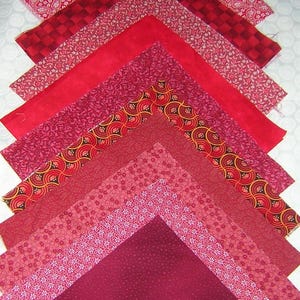 May include: An assortment of red fabric squares arranged in a chevron pattern. The fabrics display various patterns, including paisley, checks, floral, and solid colors. The color palette ranges from light pink to deep burgundy.