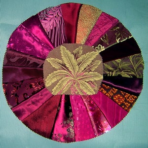May include: A circular fabric wall hanging with a central design of a stylized plant in shades of brown and gold. The surrounding fabric is arranged in a fan shape and features various shades of pink, red, and purple.