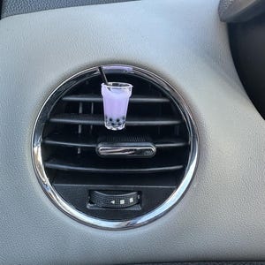 May include: A car air vent clip featuring a miniature purple bubble tea drink with a black straw. The drink is in a clear plastic cup and has dark brown boba pearls at the bottom. The air vent is silver and black.