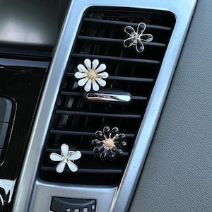May include: A car air vent featuring four flower-shaped air fresheners. The flowers are white, black, and clear, with gold and silver detailing. The vent is silver and black.