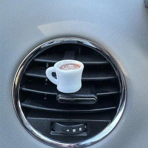 Könnte beinhalten: Ein weißer Miniatur-Kaffeebecher-Lufterfrischer für das Auto mit braunem Latte-Art-Design, platziert in einer Lüftungsöffnung des Autos. Die Lüftungsöffnung ist schwarz mit silberner Umrandung, vor einem grauen Armaturenbrett.