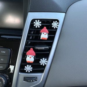 Op de afbeelding: Auto ventilatie decoraties met kleine pinguïns met rode gebreide mutsen en sneeuwvlok accenten. De pinguïns hebben witte lichamen, oranje snavels en zwarte ogen. De sneeuwvlokken zijn zilver en glinsterend.