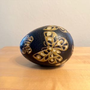 May include: A decorative, egg-shaped object with a dark blue background and intricate butterfly designs in a tan color. The butterflies are symmetrically placed around the egg's surface, creating a visually appealing pattern. The object is displayed on a wooden surface.