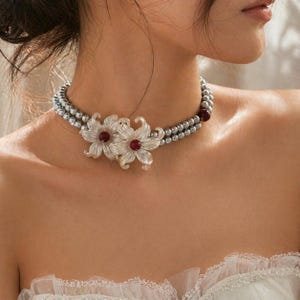 Op de afbeelding: Een ketting met twee strengen met grijze kralen en twee heldere bloemhangers met rode centra. De ketting wordt om de nek gedragen en toont het delicate bloemendesign.