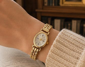 May include: A gold-tone analog wristwatch with an oval-shaped case and a white dial featuring black Roman numeral hour markers. The watch has a polished metal link bracelet and a small crown on the side. The dial includes the text Quartz.