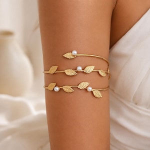 May include: Gold-toned arm cuff jewelry featuring a leaf design with small, round, white pearls. The cuff wraps around the arm, with three bands adorned with textured leaves and pearls. The jewelry is displayed on an arm against a soft, neutral background.