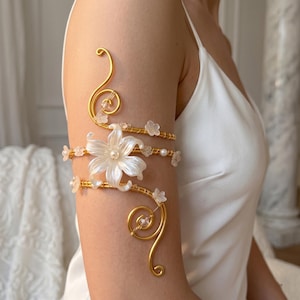 May include: Gold-toned arm cuff featuring a large white flower with a pearl center and smaller white flowers. The cuff is made of wire and has a decorative swirl design. The arm cuff is worn on the upper arm.