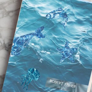 May include: A hardcover notebook with a blue ocean wave design and aquatic animal illustrations. The notebook has an orange bookmark and the text "ROBERT FISHER 2024". A cup of coffee, a pen, and a potted plant are also visible.