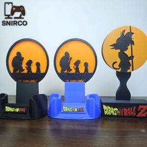 Può includere: Tre supporti per telefono a tema Dragon Ball Z. Ogni supporto presenta una silhouette nera su sfondo arancione. I supporti sono neri e blu, con la scritta "DRAGON BALL" e "DRAGON BALL Z". Il logo SNIRCO si trova in alto a sinistra.