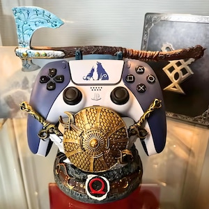 May include: A custom PlayStation 5 controller with a matching stand, featuring a God of War theme. The controller is blue and white with a wolf design, and the stand includes a shield, axe, and decorative elements. The controller is approximately 6 inches wide.