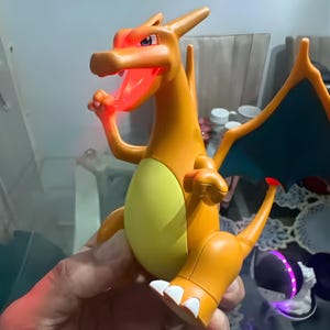 May include: Orange and yellow Charizard figure with a glowing red mouth and accents. The figure has blue wings and a yellow belly. The figure is a collectible item.