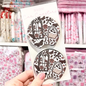 May include: Two car coasters with a white background and a repeating pattern of brown roses, butterflies, and arched window designs. The text "Car Coasters" is printed on the packaging.