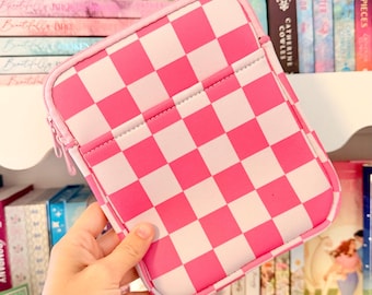 Puede incluir: Una funda protectora rectangular para tableta o dispositivo electrónico con un patrón de cuadros rosa y blanco vibrante. El estuche está hecho de un material suave con una cremallera rosa a juego y un compartimento de bolsillo frontal.