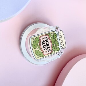 May include: A clear sticker depicting a jar of pickles with the text "PICKLE SLUT" on a pink and white banner. The jar contains green pickles and has a white lid. The sticker is on a clear backing.