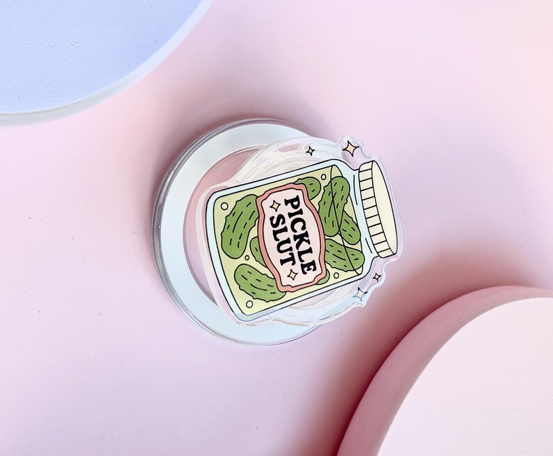 May include: A clear sticker depicting a jar of pickles with the text "PICKLE SLUT" on a pink and white banner. The jar contains green pickles and has a white lid. The sticker is on a clear backing.