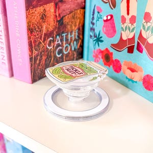 May include: A clear phone grip with a white ring base and the text "Pickle Slut". The grip is on a white shelf with books in the background. The books have pink, blue, and brown covers.