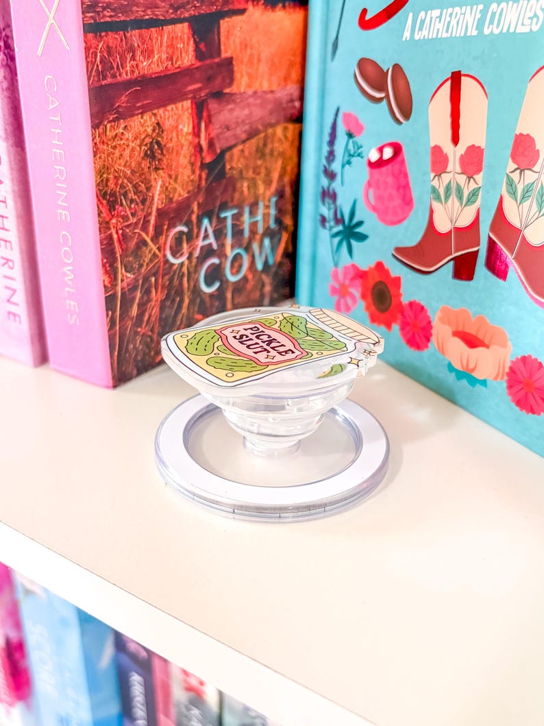 May include: A clear phone grip with a white ring base and the text "Pickle Slut". The grip is on a white shelf with books in the background. The books have pink, blue, and brown covers.