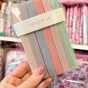 May include: A set of six pastel-colored highlighters in a clear plastic package. The highlighters are in shades of pink, gray, and green. The text "Highlighter set" is printed on the packaging. Each highlighter has the text "The Blush Hut" printed on it.