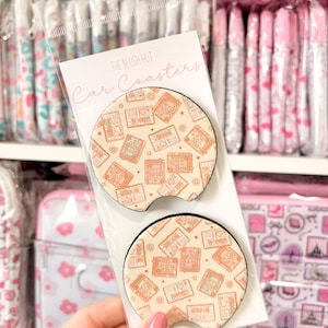 May include: Two car coasters with a book-themed design are displayed in a clear plastic package. The coasters have a beige background with a pattern of books, hearts, and stars in shades of peach and brown. The packaging reads "The Blush Hut Car Coasters."