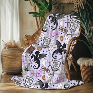 May include: A light purple throw blanket with a repeating pattern of black dragons, swords, books, feathers, and the text "BASGIATH WAR COLLEGE". The blanket is draped over a wicker chair, creating a cozy and whimsical aesthetic.