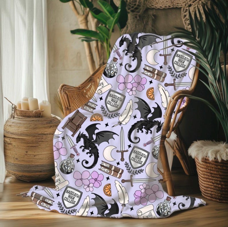 May include: A light purple throw blanket with a repeating pattern of black dragons, swords, books, feathers, and the text "BASGIATH WAR COLLEGE". The blanket is draped over a wicker chair, creating a cozy and whimsical aesthetic.