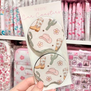 May include: Two car coasters with a western-themed design. The coasters feature a light background with illustrations of cowboy boots, cacti, cowboy hats, and flowers. The text "The Blush Hut Car Coasters" is printed on the packaging.