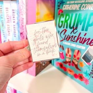 May include: A clear acrylic library card-shaped bookmark with the quote "for the girls who found themselves in a book." The bookmark is held in front of a bookshelf with books and a book titled "Grump & Sunshine."