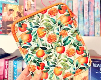 Puede incluir: Una funda con cremallera con un vibrante estampado de naranjas y hojas verdes sobre un fondo blanco. La funda tiene un ribete y una cremallera naranjas. El diseño presenta naranjas enteras y en rodajas. La funda se sostiene frente a una estantería.