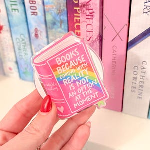 May include: A pink book-shaped phone grip with the text "BOOKS BECAUSE COPING WITH REALITY IS NOT AN OPTION AT THE MOMENT" and a rainbow design. The grip is held in front of a shelf of books.
