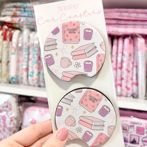 May include: Two car coasters with a white background and a pattern of pink books, candles, mugs, and flowers. The coasters are packaged in a clear plastic sleeve. The text "Bookish Girls" is visible on one of the books.