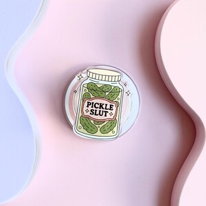 May include: A round sticker showing a jar of green pickles with the text "PICKLE SLUT" on a label. The jar has a white lid and is set against a light pink and blue background. The sticker has a clear border.