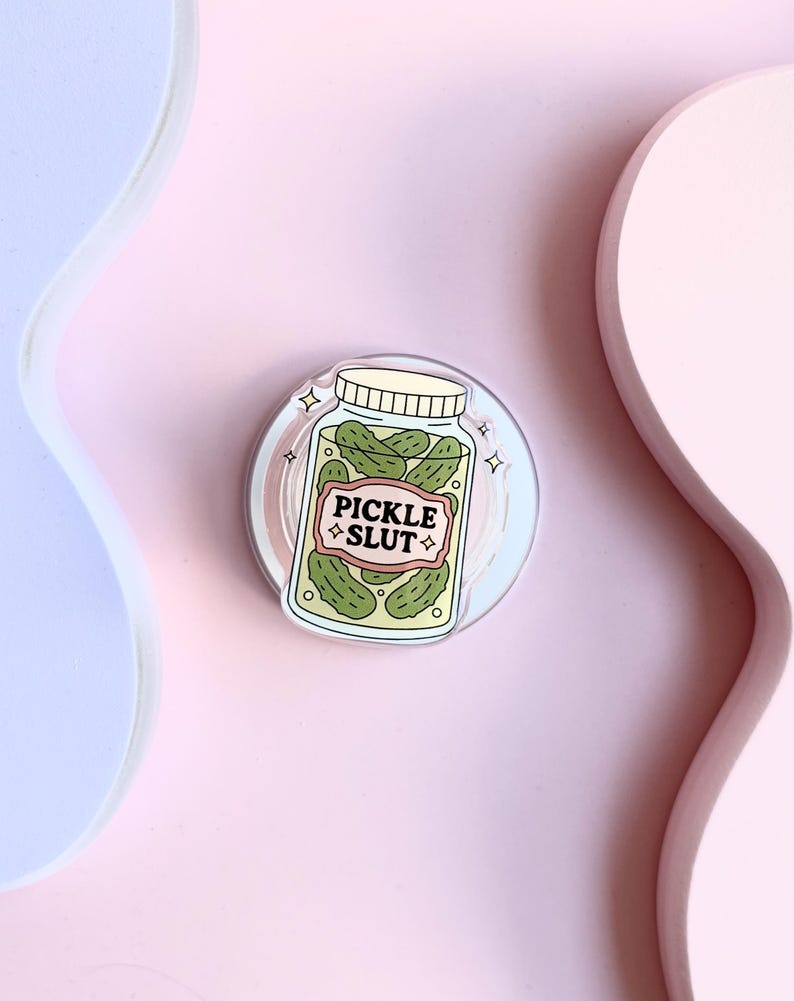May include: A round sticker showing a jar of green pickles with the text "PICKLE SLUT" on a label. The jar has a white lid and is set against a light pink and blue background. The sticker has a clear border.