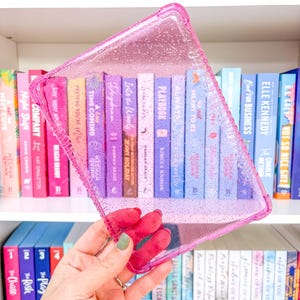 Puede incluir: Una funda transparente y rosa brillante para libros se sostiene frente a una estantería. La funda es rectangular con un borde elevado. Los libros tienen varios colores y títulos, incluyendo "Playbook" y "Always Meant to Be".