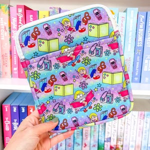 Può includere: Una colorata custodia per libri con un motivo ripetuto di una ragazza che legge, libri, telefoni cellulari, pretzel, stelle e fiori. La custodia ha una cerniera e un bordo bianchi. Lo sfondo è un mix di blu, viola e rosa.