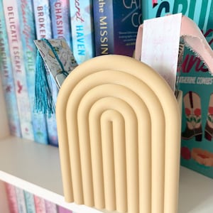 Puede incluir: Un sujetalibros beige arqueado con diseño de arcoíris sobre una estantería blanca. El sujetalibros está frente a una fila de libros con cubiertas coloridas. Se ve un marcapáginas con una borla azul.