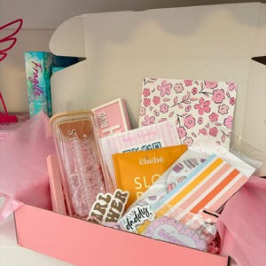 Puede incluir: Una caja rosa llena de varios artículos, incluyendo un frasco de vidrio con tapa de madera, un lápiz labial rosa, pegatinas y un cuaderno con estampado floral. La caja está forrada con papel de seda rosa y contiene el texto "GIRL POWER".