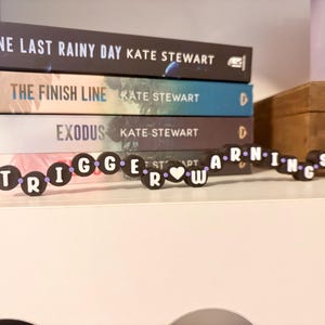 May include: A stack of books with titles including "One Last Rainy Day," "The Finish Line," and "Exodus." A decorative word chain spells out "Trigger Warnings" in black and white. The books are on a white shelf.