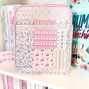 May include: A patchwork tablet case with a pink zip and trim. The case showcases floral and cherry patterns in pink, white, and grey. It's displayed on a white shelf with books in the background, creating a charming aesthetic.