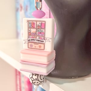 May include: A charm featuring a stack of miniature books and a white block with a bookshelf design and the text "NO SHELF CONTROL." The charm is attached to a keychain with a pink bead and a black and white patterned bead.