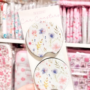 May include: Two car coasters with a floral design in a clear plastic package. The coasters have a white background with pink, blue, and yellow flowers and green leaves. The text "THE BLUSH HUT Car Coasters" is printed on the packaging.