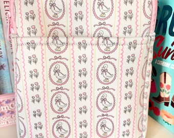 Puede incluir: Una funda para libros estampada en rosa y blanco, decorada con patos en marcos ovalados, lazos y motivos florales. La funda tiene una cremallera y un ribete rosas. La funda para libros está en una estantería con libros.