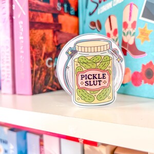 May include: A clear sticker depicting a jar of pickles with the text "PICKLE SLUT" on a pink and yellow label. The sticker is on a white shelf, with colourful books in the background. The jar contains green pickles.