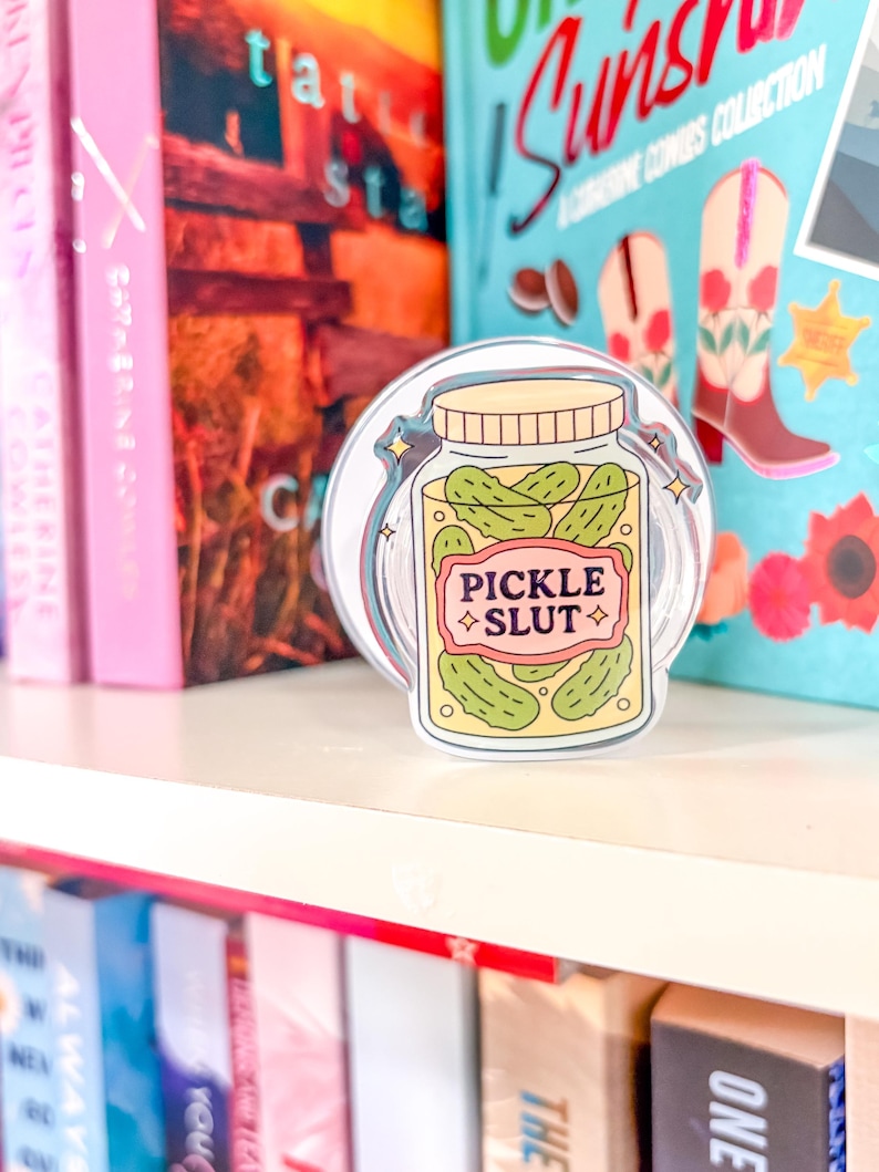 May include: A clear sticker depicting a jar of pickles with the text "PICKLE SLUT" on a pink and yellow label. The sticker is on a white shelf, with colourful books in the background. The jar contains green pickles.