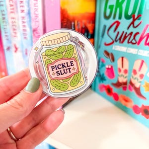 May include: A clear, round phone grip featuring a cartoon jar of pickles with the text "PICKLE SLUT" on a pink label. The grip is held in front of a bookshelf with colorful book spines.