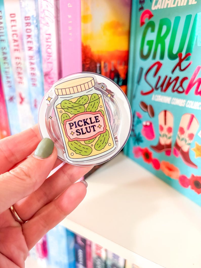May include: A clear phone grip with a cartoon jar of pickles and the text "PICKLE SLUT" on a pink label. The grip is held in front of a bookshelf with colorful book spines.